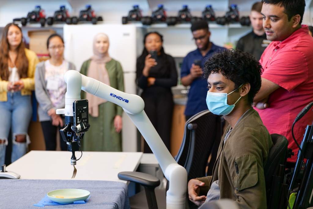 The students and instructor gather around a white robotic arm, which holds a fork above a plate with food on it